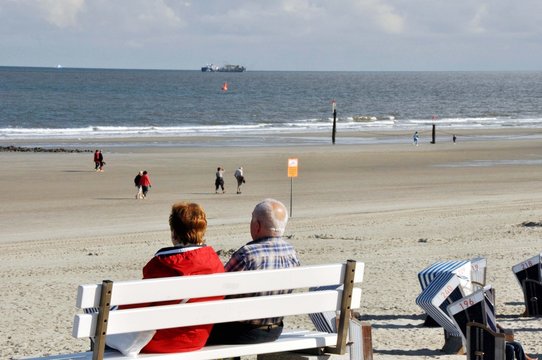Ebbe an der Nordsee / ideal f&uuml;r lange entspannte Strandspazierg&auml;nge. Ein &auml;lteres Paar verweilt auf einer Bank oben an der Strandpromenade mit Blick auf das weite Meer mit Frachter