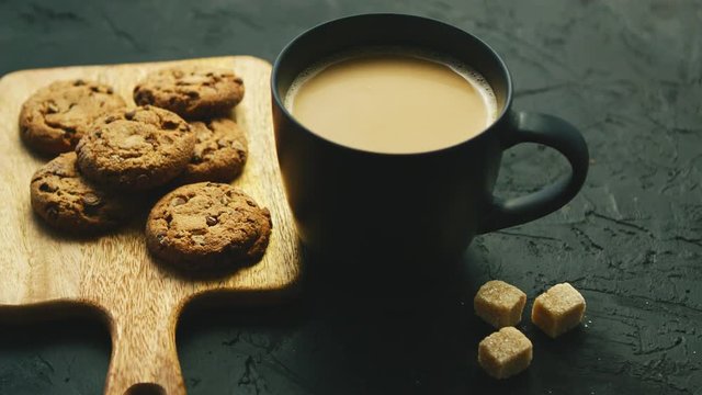 From above view of mug of hot coffee with biscuits laid on wooden cutting board and pieces of brown sugar on gray background