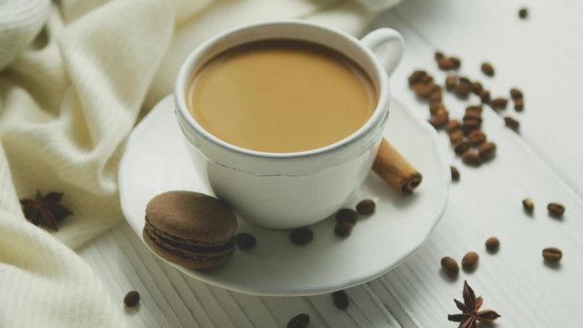 From above view of cup of coffee with biscuit and cinnamon decorated with beans on white wooden background