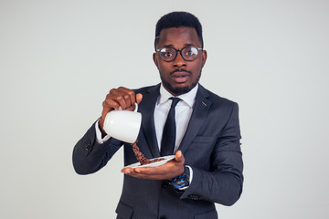 surprised and amazed handsome afro american business man in a black classic suit and glasses holding a cup with flying coffee bean splash on plate in white background studio shot. Magic morning drink