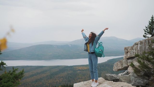 Attractive woman standing on a mountain peak with her long hair blowing in the wind smiling and trying to keep her hair off her face with her hand