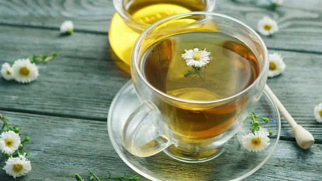 Closeup shot of glass mug on saucer filled with herbal tea and tiny camomile flowers on wooden table with jar of honey