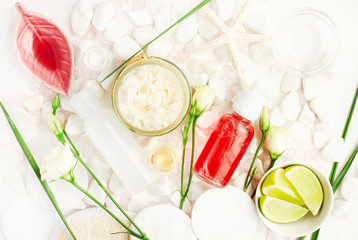 Skincare scrub with Coconut flakes in glass jar, red holistic facial lotion bottle, fresh lime and flowers viewed above on white spa stones. 