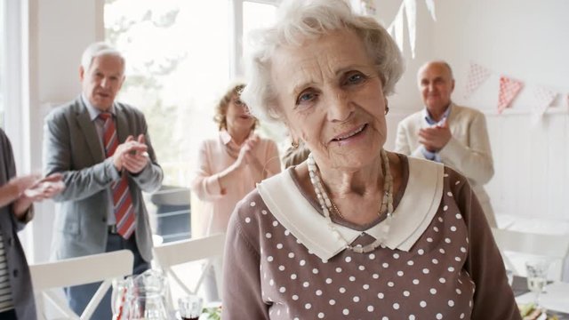 Portrait of happy senior woman smiling and posing for camera in the living room while joyous friends standing in background and clapping hands at birthday party