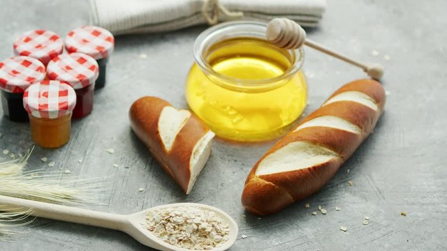 Composition of fresh baguettes with jar of honey on table near spoon of oats and small containers with marmalade