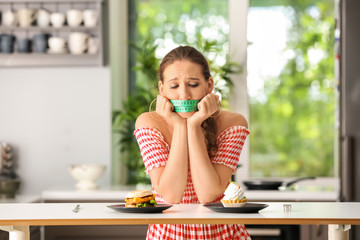 Emotional woman with measuring tape around her mouth and unhealthy food in kitchen. Diet concept