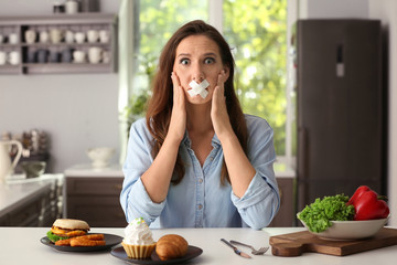 Stressful woman with taped mouth and different products in kitchen. Choice between healthy and...