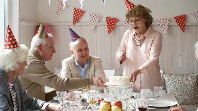 Cheerful senior woman in party hat smiling and chatting with guests while cutting birthday cake at dinner party