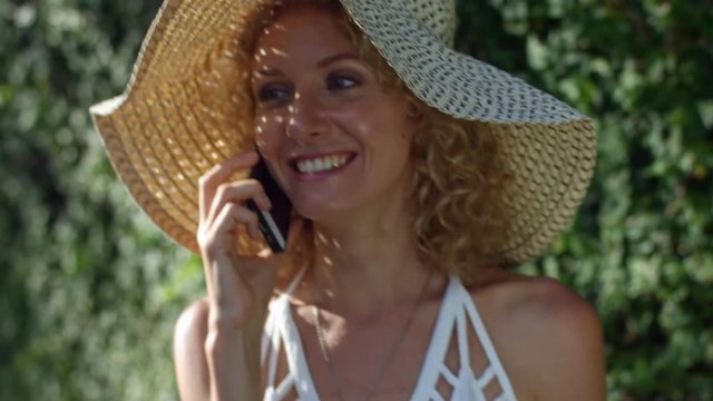 Medium shot of young blonde woman in straw hat smiling when having phone conversation outdoors