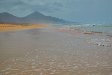 Beautiful paradisiacal beach with golden sand and waves with the mountain in the background and with clouds in Cofete, Fuerteventura, Canary Islands, Spain