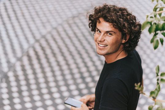 Rear view image of cheerful man in black t-shirt reading text message on his mobile phone on the street. Young Caucasian male with curly hair looking to the camera with copy space for text.