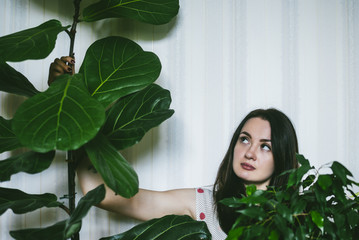 Fototapeta premium Pretty brunette girl sitting near flowerpot on the floor of the living room and looking in the distance. Cozy leisure atmosphere at home.