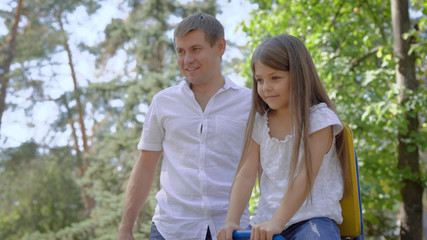 Fototapeta premium loving father is helping to his little daughter to sway on swing in playground in summer day, girl is smiling