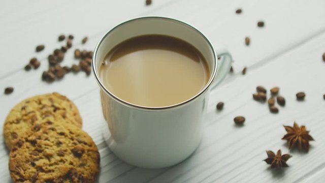 White cup with coffee and milk on white wooden table with cookies and coffee grains on top around