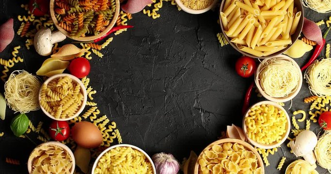 From above shot of bowl with different pasta and macaroni composed in circle with cooking ingredients on black table