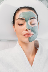 overhead view of beautiful woman with eyes closed and clay mask on face in white bathrobe relaxing in spa salon