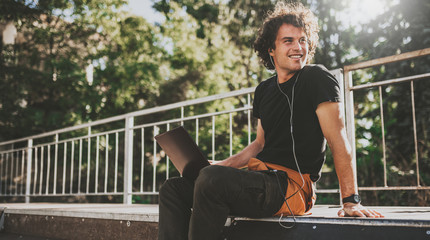 Handsome male with curly hair using laptop computer chatting online with friends using earphones and laptop connected to free wireless. Cheerful male blogger publishing post on website on computer.