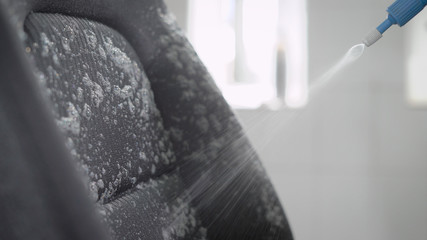 washer is spraying foam on a upholstery of seats inside automobile and rubbing by brush, in a garage, close-up
