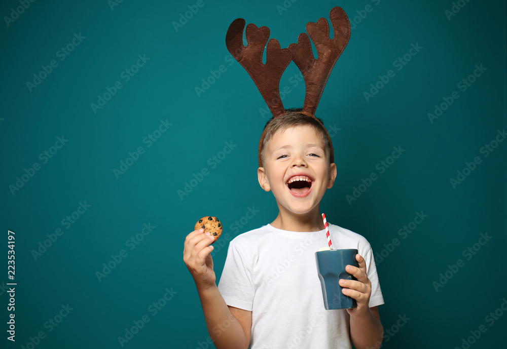 Wall mural cute little boy with toy reindeer horns holding cup of hot chocolate and cookie on color background