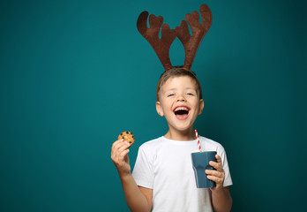 Cute little boy with toy reindeer horns holding cup of hot chocolate and cookie on color background © Pixel-Shot