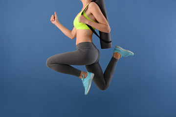 Young woman with yoga mat jumping on color background