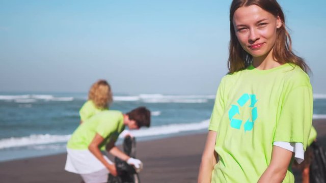 Four volunteers in green t-shirts with image recycle collect garbage on beach, looking on camera with bags of collected garbage. Volunteering and recycling concept. Environmental awareness copy space