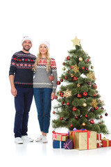 Obraz na płótnie Canvas beautiful happy couple in santa hats standing near christmas tree with gifts, isolated on white