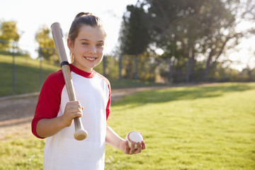 Young girl holding baseball and baseball bat looks to camera