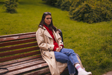 Portrait of woman in full length wearing beige trench coat, red hoodie, blue jeans while walking in park and sitting on bench.