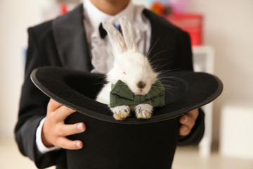 Cute little magician holding hat with rabbit indoors, closeup