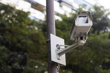 Bellow view of a security camera(cctv) in a public place. With a metal camera, green trees and buildings in the background we compose this photo. Secure, systens, technology and monitoring as concept