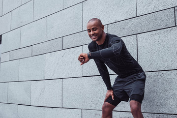 Ready and go. Young man in sportswear looking at his smartwatch with smile while standing against grey background outdoors