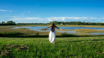 Back of a Brunette young woman bride with long curly hair wearing white walking arms extended embracing eart and sky in a landscape next to a lake during the sunset