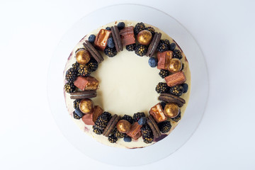 Cake with white and black cream, decorated with blueberries, blackberry, chocolate chip cookies, candies, bars and gold on a white background. Top view.