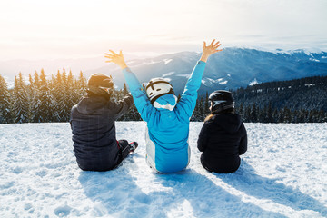 Three people in ski suits are sitting on the snow at the top of the mountain and looking into the...