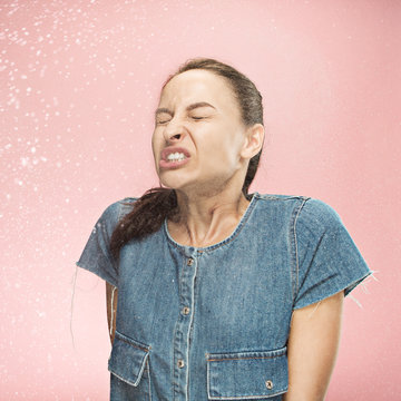 Young funny woman sneezing with spray and small drops, studio portrait on pink background. Comic, caricature, humor. illness, infection, ache. Health concept