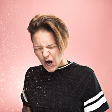 Young funny woman sneezing with spray and small drops, studio portrait on pink background. Comic, caricature, humor. illness, infection, ache. Health concept