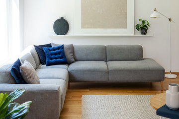 Blue pillows on grey corner sofa in apartment interior with lamp and plant next to poster. Real photo