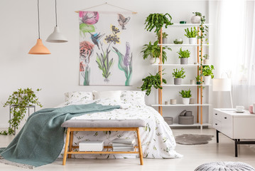 Close to nature bright bedroom interior with a bed covered with white sheets and marine blanket. Green plants on shelves next to the bed. Real photo. - obrazy, fototapety, plakaty