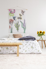 Cottage house minimal bedroom interior with colorful flowers and birds painted on fabric above a bed which is dressed in natural textile sheets. Sunflowers by the bed. Real photo.