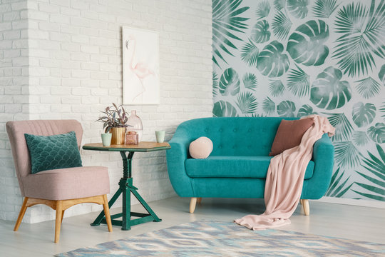 Real photo of turquoise couch and pastel pink armchair standing in bright living room interior with flamingo poster on brick wall and leafy wallpaper