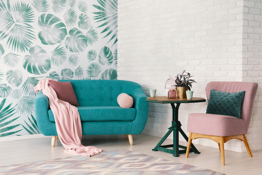 Real photo of bright sitting room interior with wooden end table with fresh plant and two mugs standing between dirty pink armchair and turquoise lounge with pillows and blanket