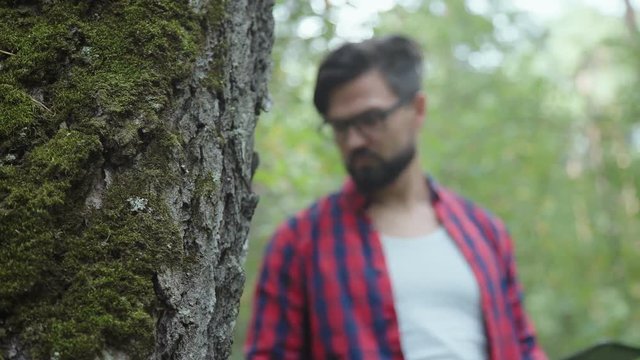 A man in a plaid shirt and glasses in the woods alone. He carefully and with interest considers a tall tree with moss on the trunk. Concept botanist, scientist, ecologist.
