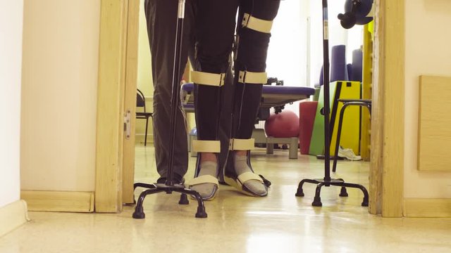 Young disable man in orthosis walking with support of two walking cane in the rehabilitation clinic. Doctor helping him.