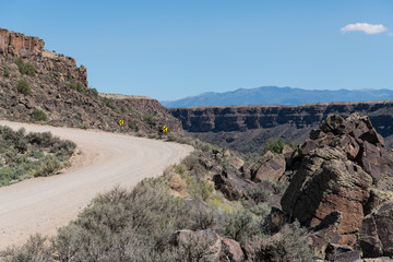 Dirt road curving up to a canyon rim with left arrow signs pointing towards a switchback - the Rio Grande Gorge near Taos, New Mexico with the Sangre de Cristo mountain range in the distance