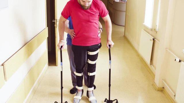 Young disable man in orthosis walking with support of two walking cane in the rehabilitation clinic. Doctor helping him.