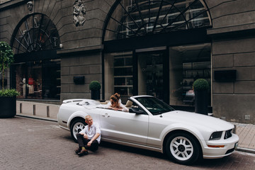 Obraz premium A newlyweds trip in the car. The groom invites the bride in a white cabriolet.