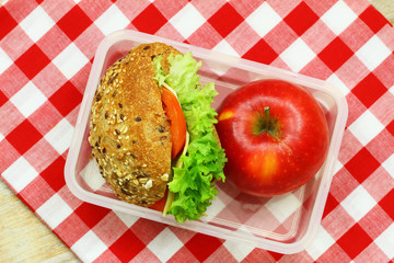 Mixed grain roll with cheese, lettuce and tomato and red apple in lunch box on red and white checkered cloth
