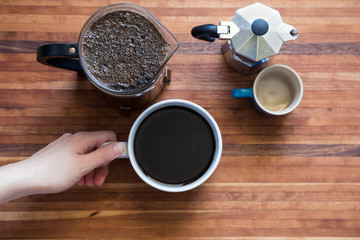 A hand grabbing the handle of a mug of coffee with a french press and smaller pot on a wood cutting board.