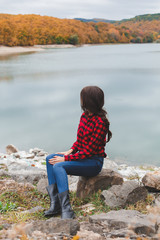 Naklejka na ściany i meble Young woman wearing red plaid shirt sitting alone near the lake in autumn. Weekend outdoors, cloudy cold weather, fall colors. Back view.